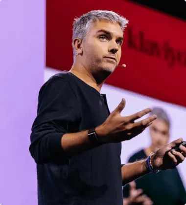 A person with a headset microphone gestures while speaking on stage, with a blurred presentation screen in the background.