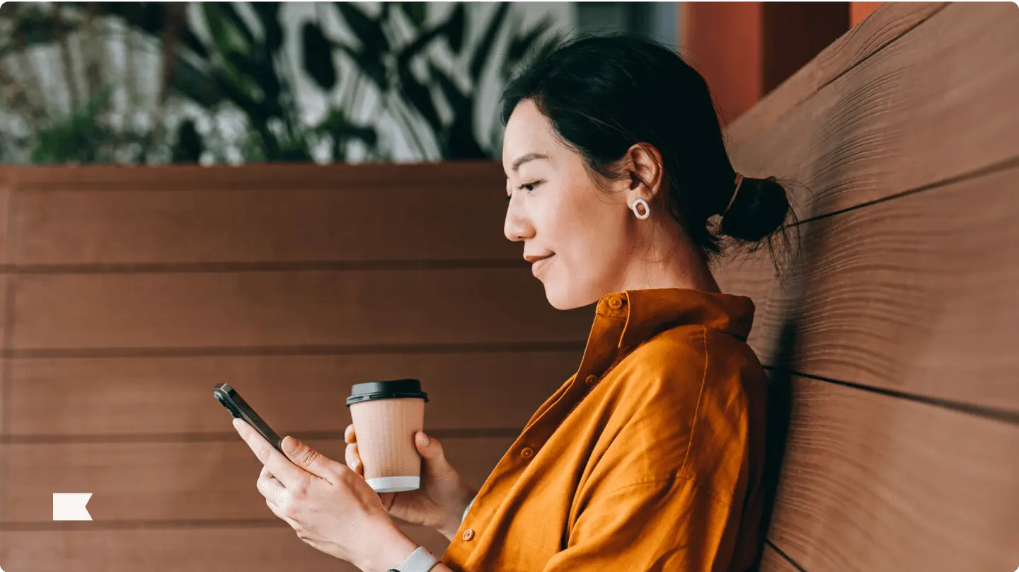 Woman in an orange shirt holding a coffee cup and smartphone, sitting on a wooden bench.