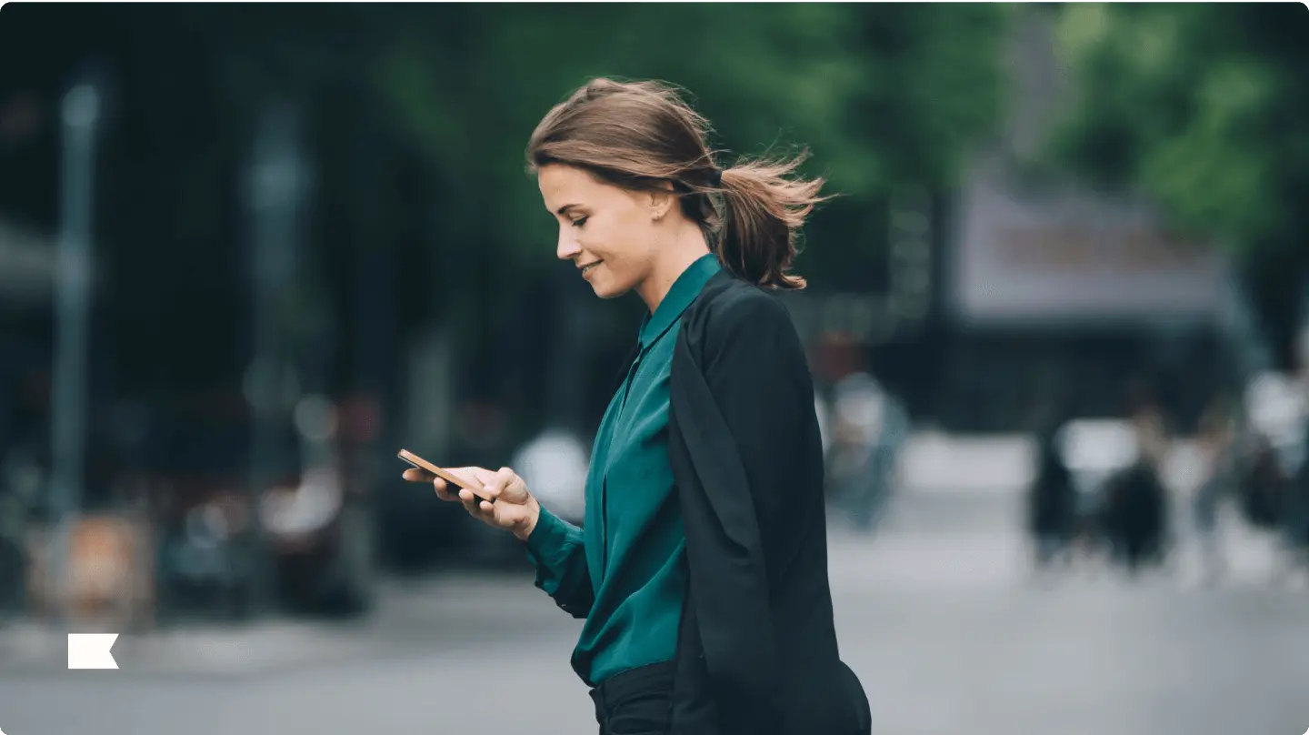 A woman in a green blouse and black jacket is walking outdoors, looking at her smartphone.