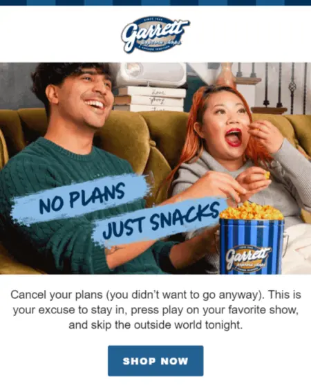  Image shows two people on a couch eating popcorn. The person on the left is a tanned-skin person with dark hair, wearing a green sweater. The person on the right is a light-skinned person with orange hair and red lipstick. Both are smiling and eating out of a blue-striped Garrett Popcorn bin.